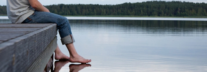 A person sits on a wooden dock with their feet touching a calm lake, surrounded by a quiet forest under a cloudy sky.