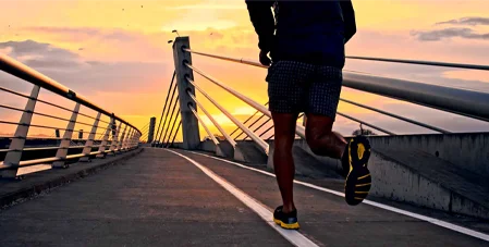 A men runs across a modern bridge at sunset, surrounded by warm orange skies and urban architecture.